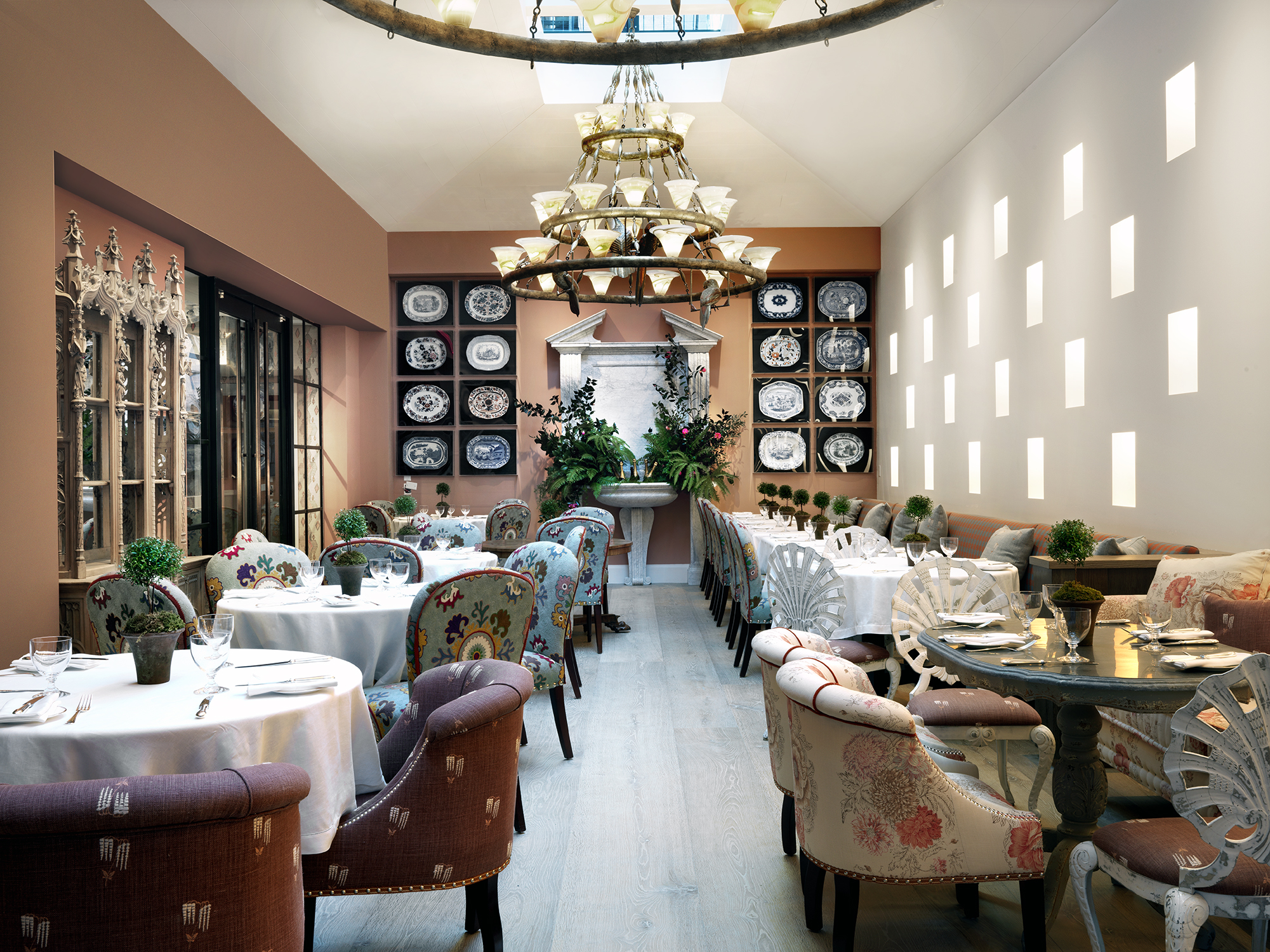 The Orangery with plenty of comfortable seats and tables for dining. A wall with sixteen art pieces showcasing various serving plates scattered around a leafy fountain oasis.