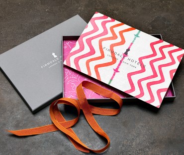 A picture of the actual voucher that will be received in the post. A grey box with Firmdale Hotels and logo applied. Inside the box is lined in a bright pink print of a Kit Kemp fabric design. The voucher is white with black writing and edged in a colourful print of rick rack designed in orange and pink. The box is tied with orange ribbon.