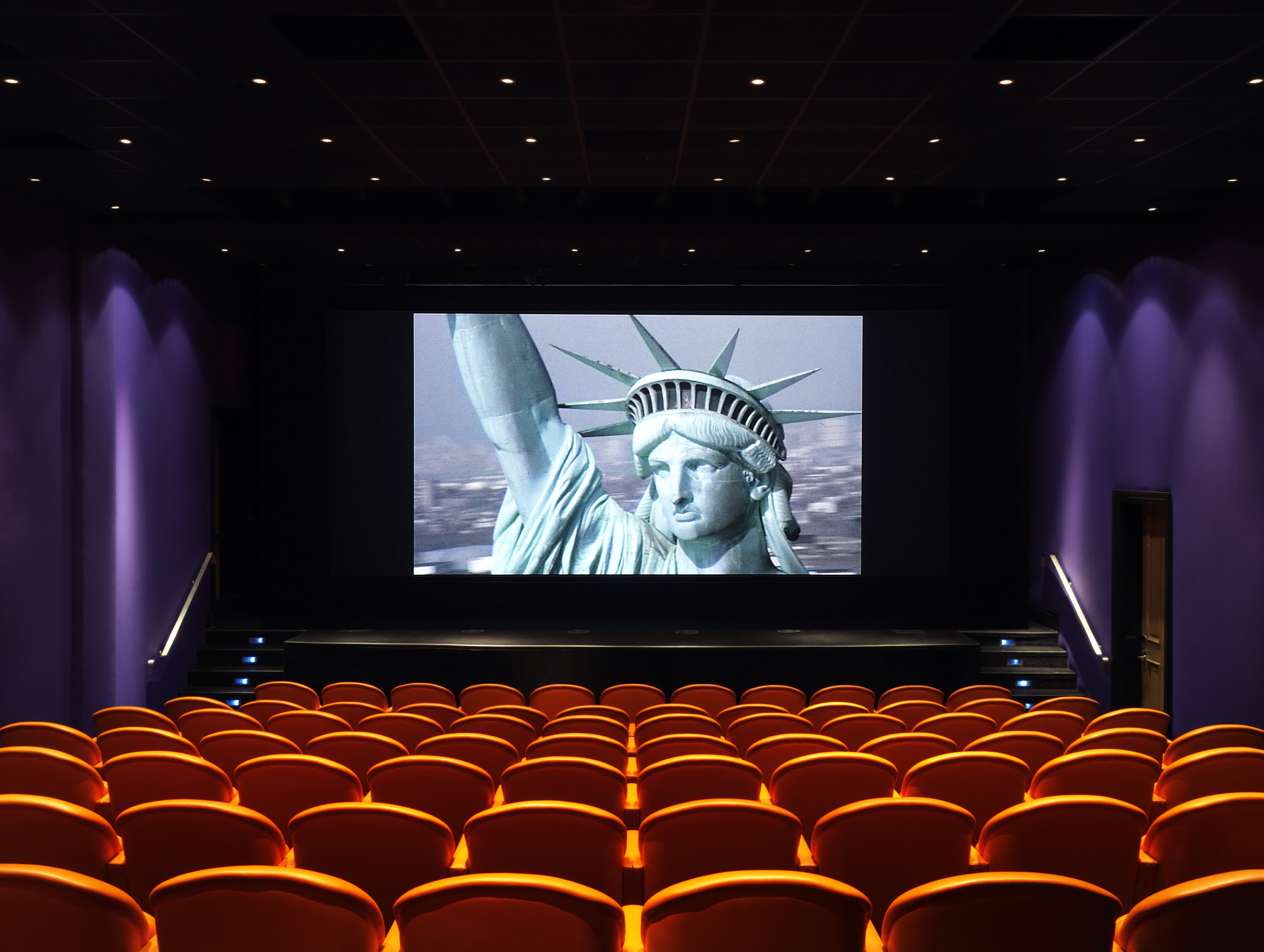 The screening room at the Crosby Street Hotel has 99 red leather seats by the Italia furniture company Poltrona Frau. there is a stage and wide screen here pictured with an image of the Statue of Liberty.