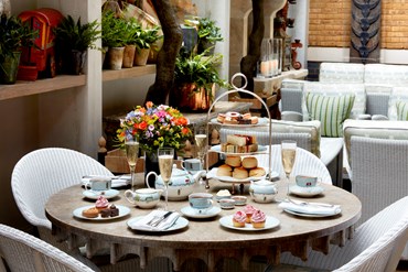 Champagne afternoon tea displayed on a table in Ham Yard Hotel. The fine bone china is Mythical Creatures by Kit Kemp for Wedgwood along side a 3 tier cake stand, sandwiches and scones.